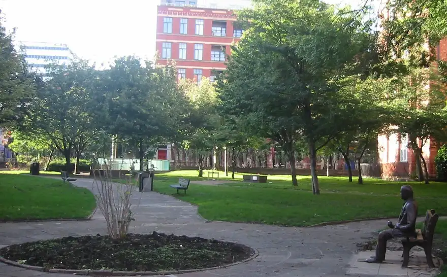 A bronze statue of Alan Turing seated on a bench in a city park, holding an apple in one hand